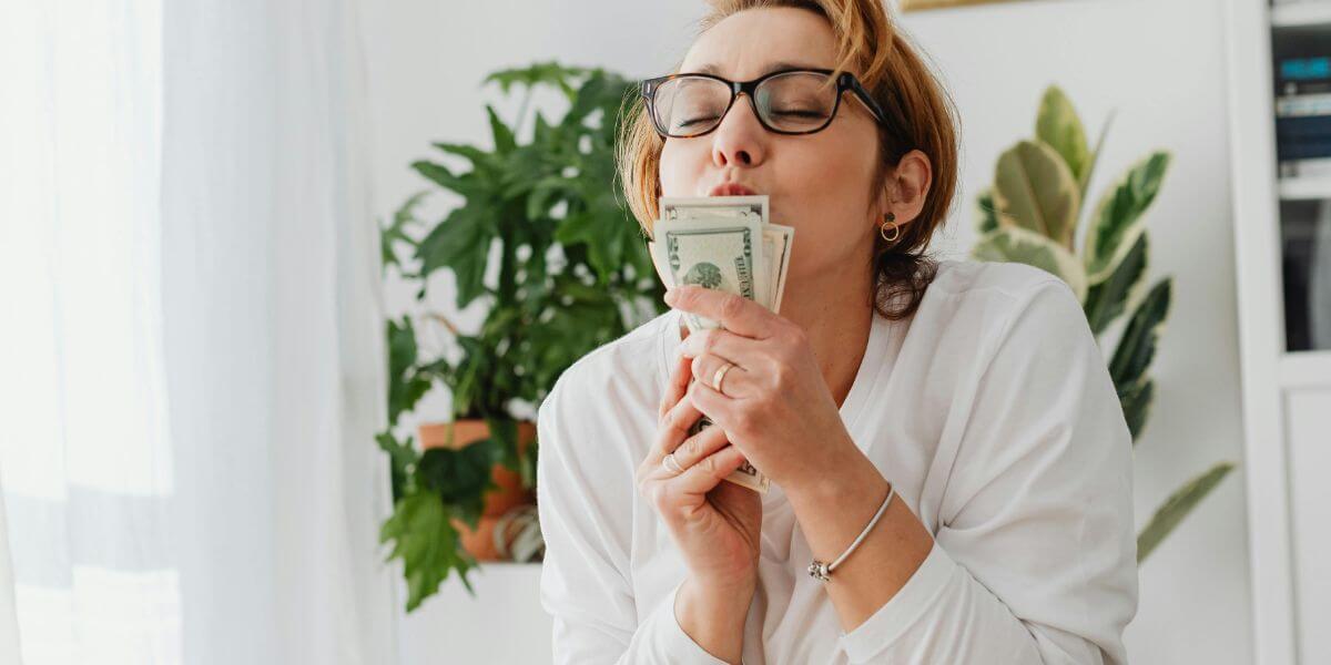 Imagem de uma mulher empreendedora, apoiada em uma mesa de escritório, beijando seu dinheiro.